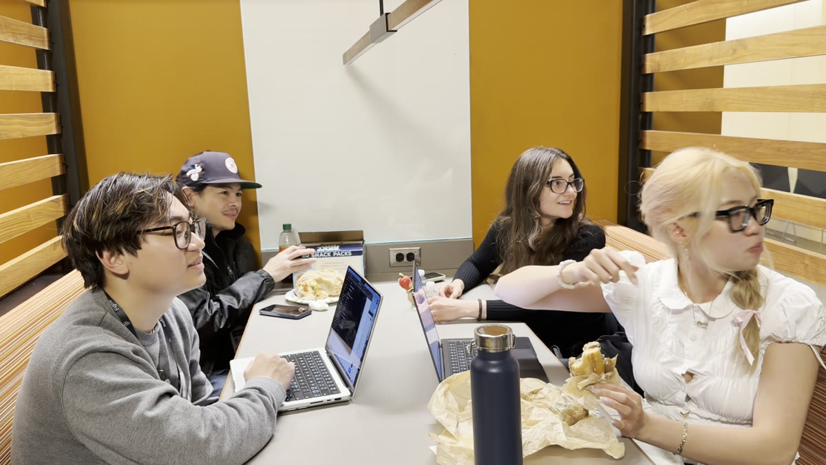 The four teammates around a table of laptops and snacks, mid-conversation while they build.